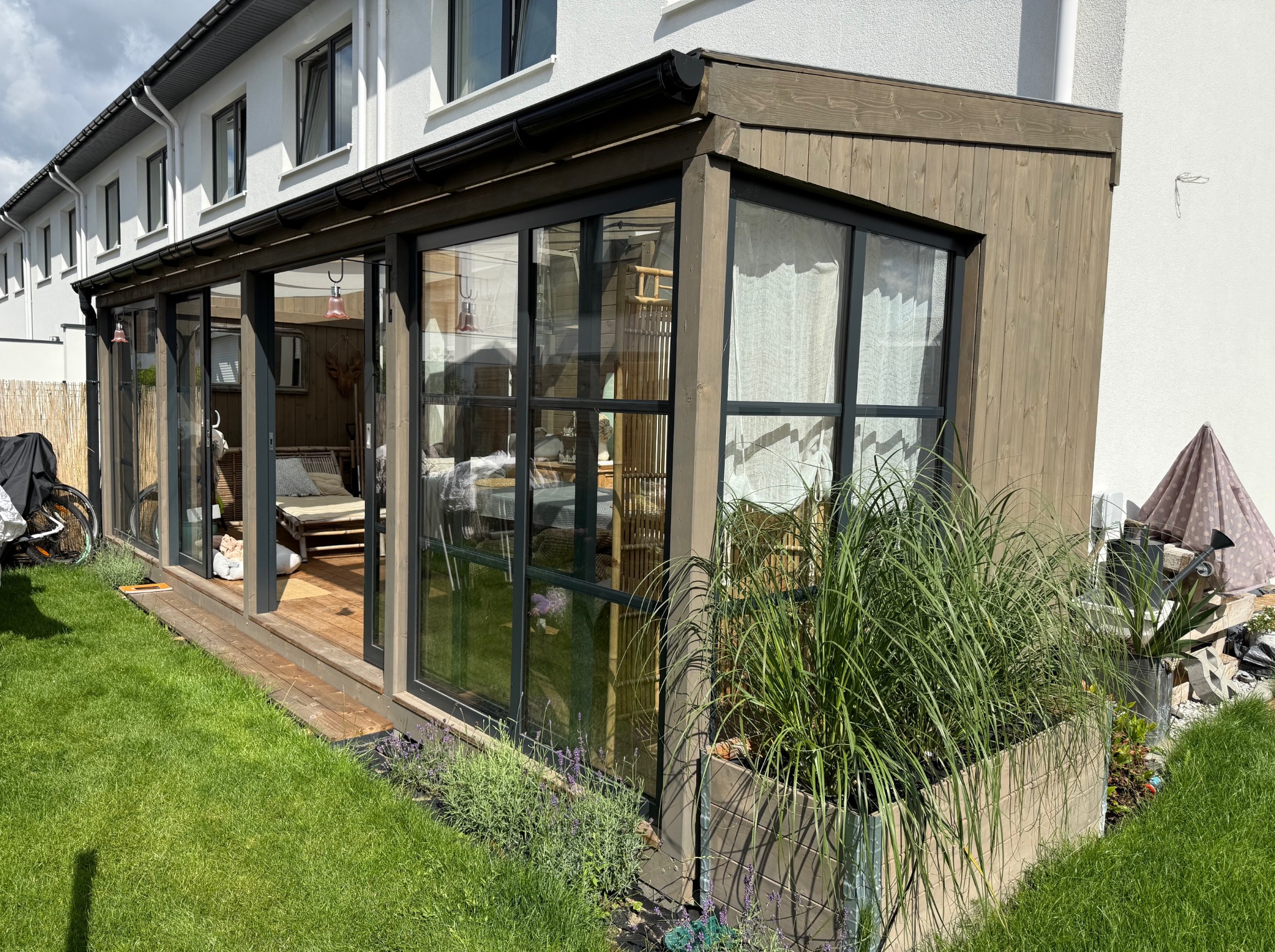 Zadaszony taras z drewnianą konstrukcją i dużymi oknami, widok z zewnątrz na trawnik i rośliny w donicach. Widoczny fragment budynku mieszkalnego.