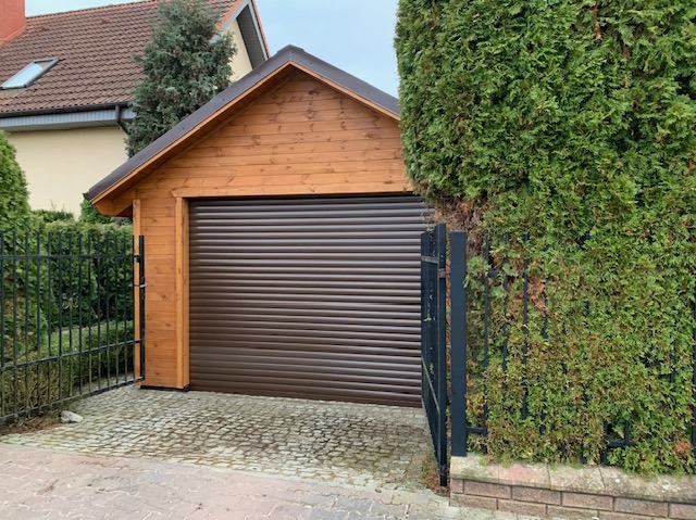 Drewniana wiata garażowa z brązową bramą rolowaną, widoczna zza czarnego metalowego ogrodzenia porośniętego zielenią.