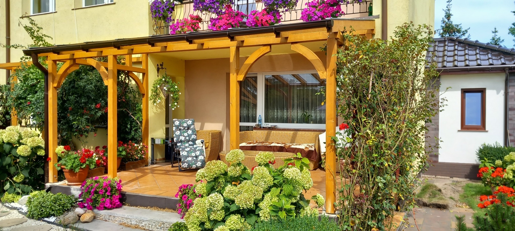 Drewniana pergola z łukowatymi podporami, zadaszająca taras z meblami ogrodowymi i kwiatami. Ujęcie z ogrodu, widok na balkon z kwiatami.