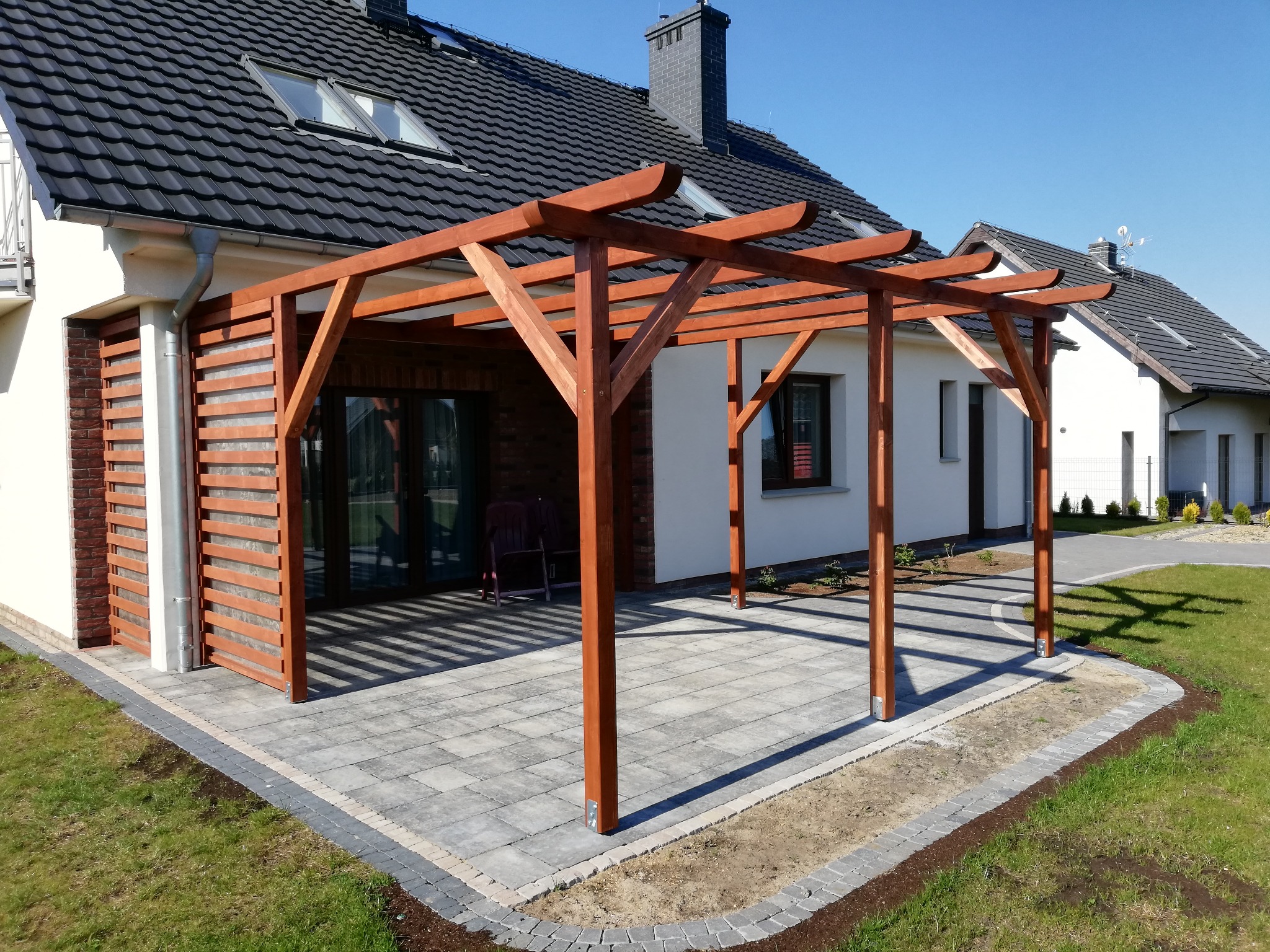 Drewniana pergola przy domu z kostką brukową i trawnikiem w słoneczny dzień. Widoczne elementy konstrukcyjne i detale wykończenia.