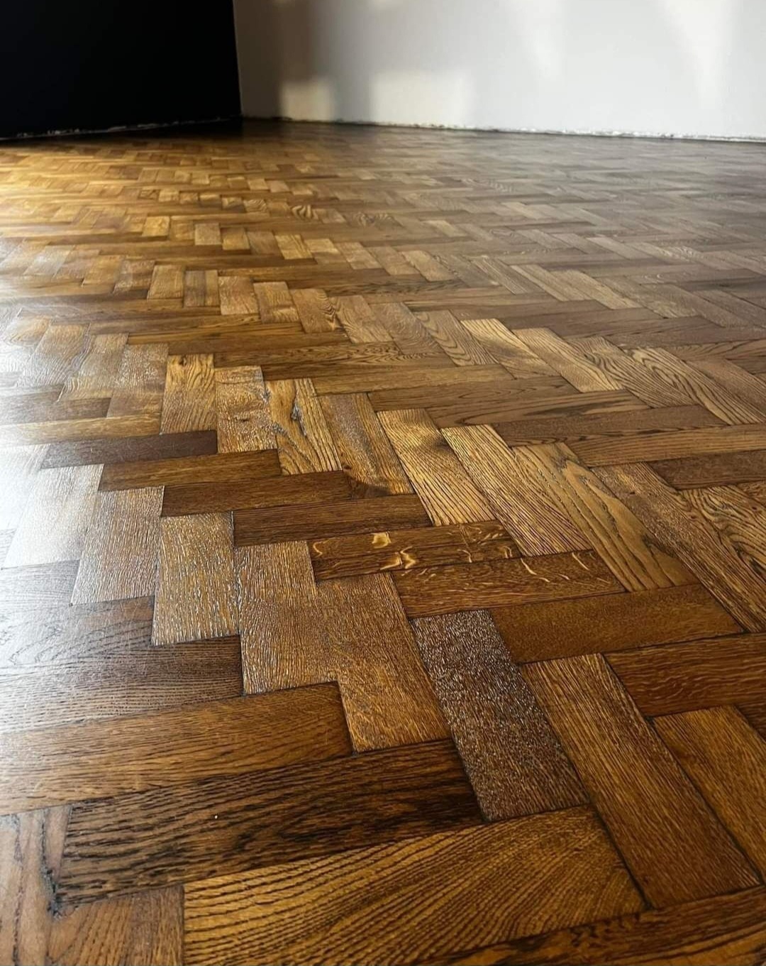 Ułożona w jodełkę, lakierowana podłoga z parkietu dębowego w odcieniach brązu, widok z niskiego kąta, z fragmentem białej ściany i czarnych drzwi w tle.