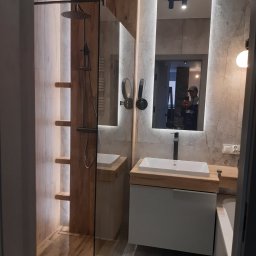 Wykończenia wnętrz Łódź - Nowoczesna łazienka z kabiną prysznicową typu walk-in, drewnopodobnymi płytkami, oświetleniem LED i dużym lustrem z podświetleniem, w którym odbija się osoba robiąca zdjęcie.