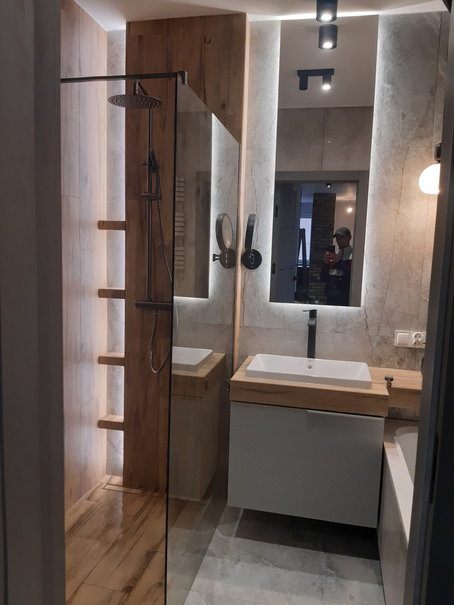 Nowoczesna łazienka z kabiną prysznicową typu walk-in, drewnopodobnymi płytkami, oświetleniem LED i dużym lustrem z podświetleniem, w którym odbija się osoba robiąca zdjęcie.