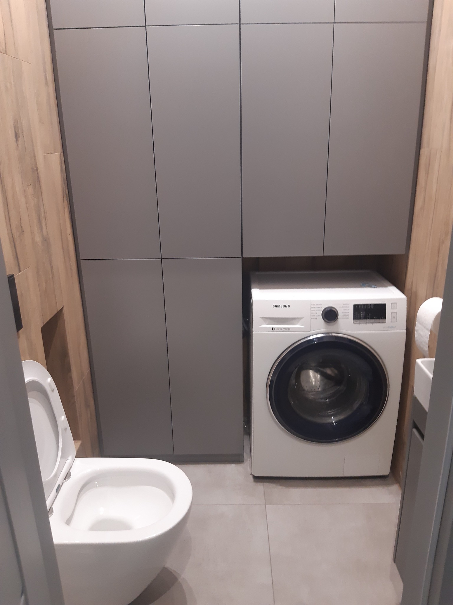Mała łazienka z szarą zabudową meblową, białą pralką Samsung i toaletą z otwartą klapą.