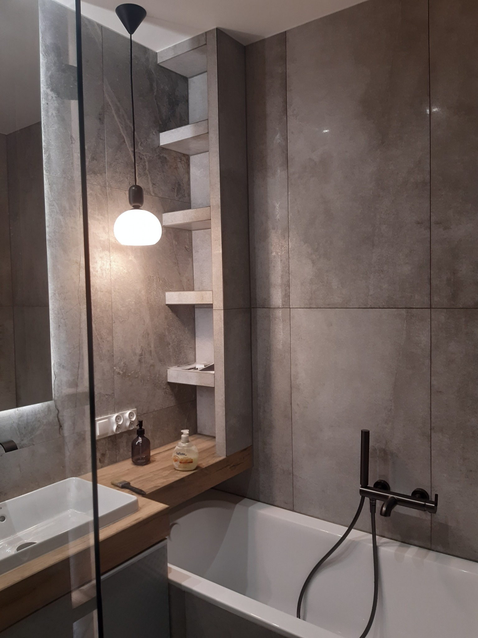 Szara łazienka z płytkami imitującymi kamień, biała wanna, umywalka nablatowa na blacie z drewnopodobnego materiału, wisząca lampa z okrągłym kloszem i półki w zabudowie.