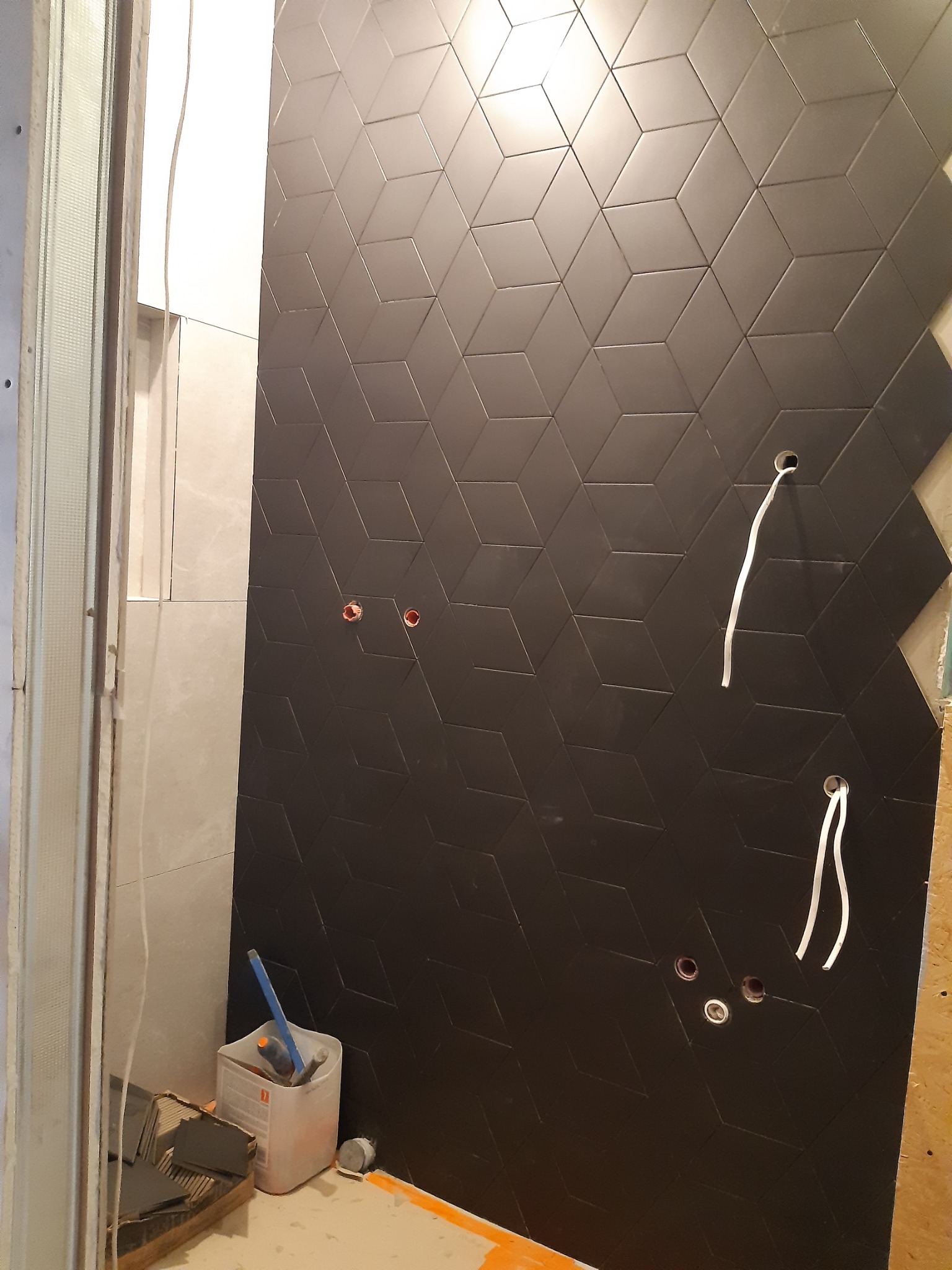 Montaż czarnych, sześciennych płytek na ścianie w łazience, widoczne otwory na instalacje, obok narzędzia i materiały budowlane.