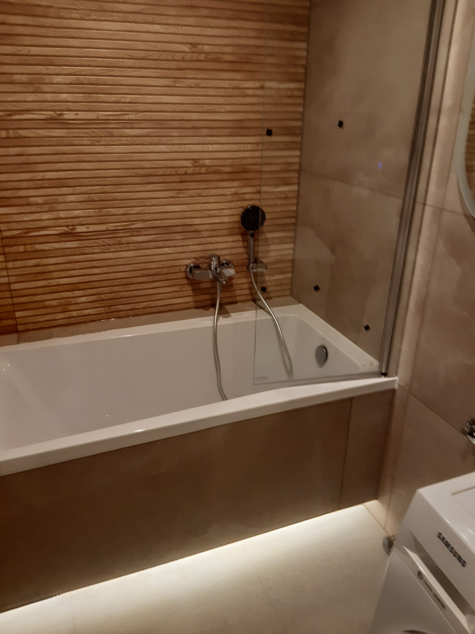 Nowoczesna łazienka z wanną obudowaną brązowymi panelami, szklanym parawanem nawannowym i drewnopodobną okładziną ścienną z poziomych listew.