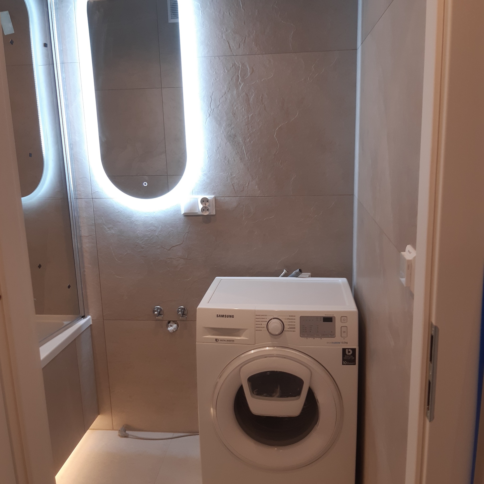 Nowoczesna łazienka z pralką Samsung, podświetlanym lustrem i beżowymi płytkami imitującymi kamień.