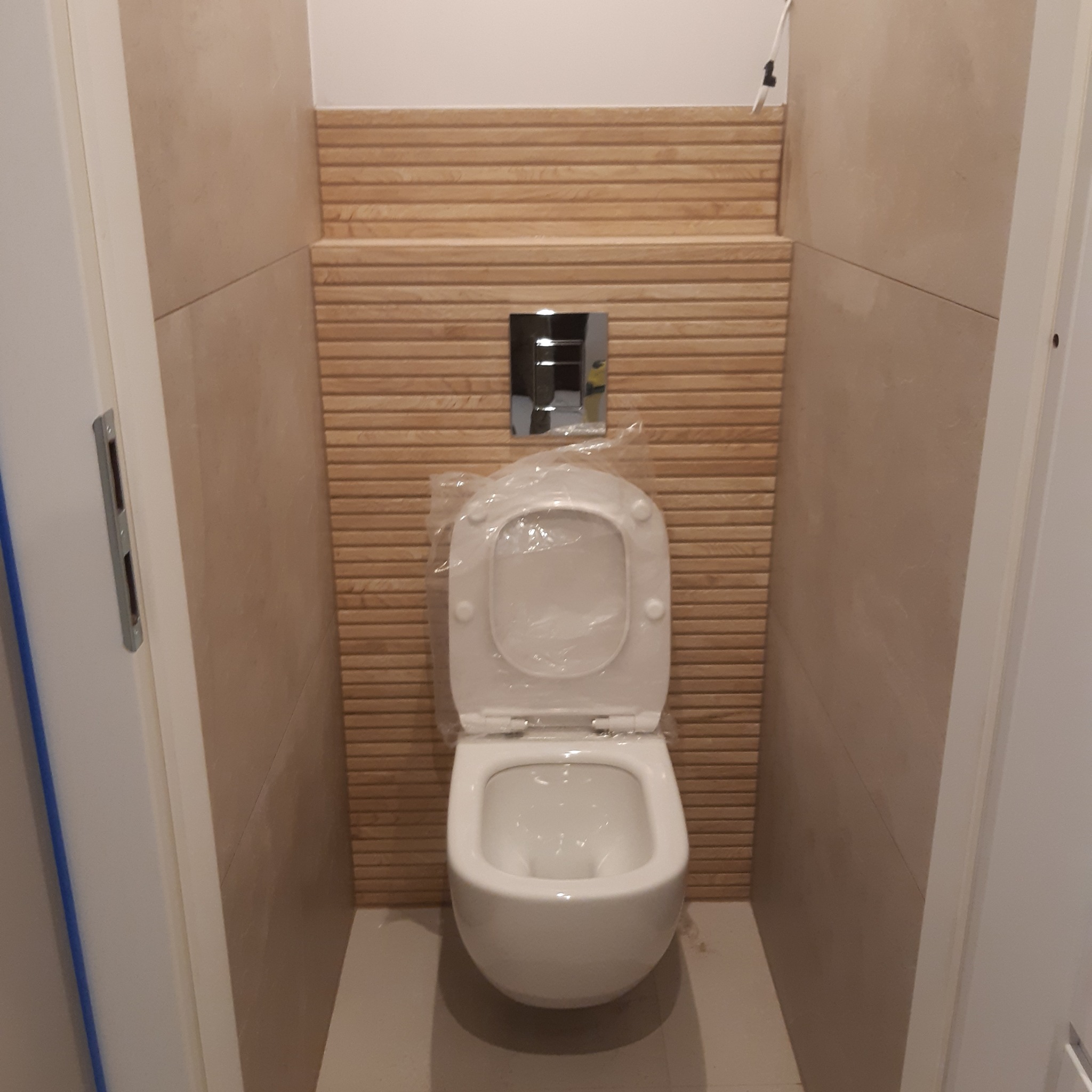 Zamontowana, biała toaleta wisząca z chromowanym przyciskiem spłukującym na tle ściany wyłożonej płytkami imitującymi drewniane lamele; sedes jeszcze w folii ochronnej, widoczne jasne płytki...