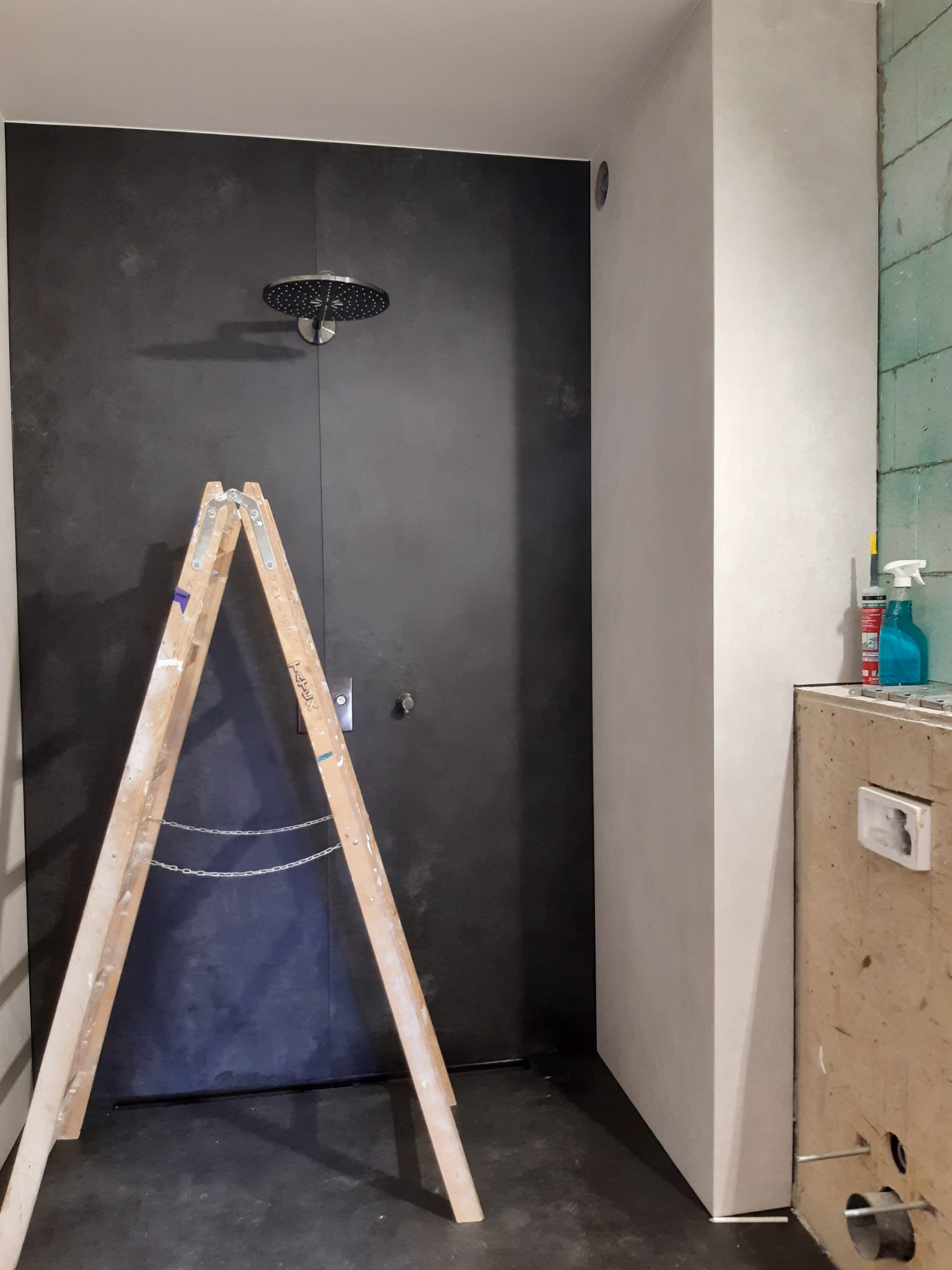 Łazienka w trakcie prac wykończeniowych: czarna ściana prysznicowa z zamontowaną deszczownicą, drewniana drabina oparta o ścianę, fragment ściany z surowym wykończeniem i widocznymi materiałami...