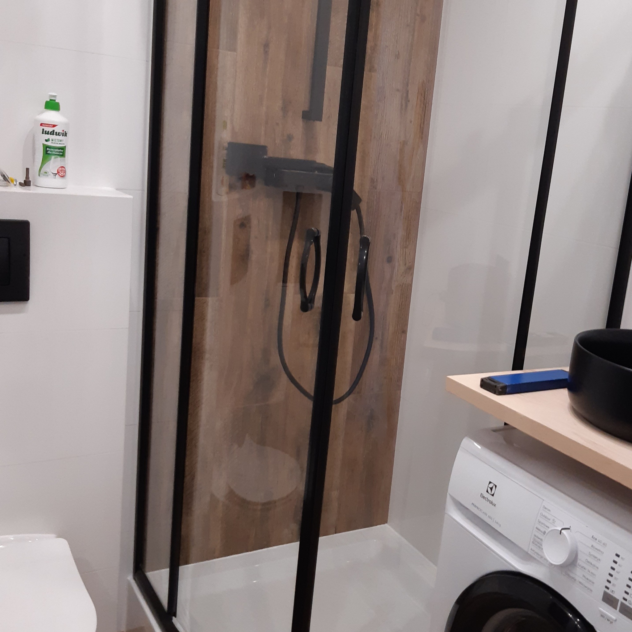 Nowoczesna kabina prysznicowa z czarnymi profilami i drewnopodobną ścianą w tle, obok umywalka nablatowa i pralka Electrolux, widoczny płyn do mycia naczyń.