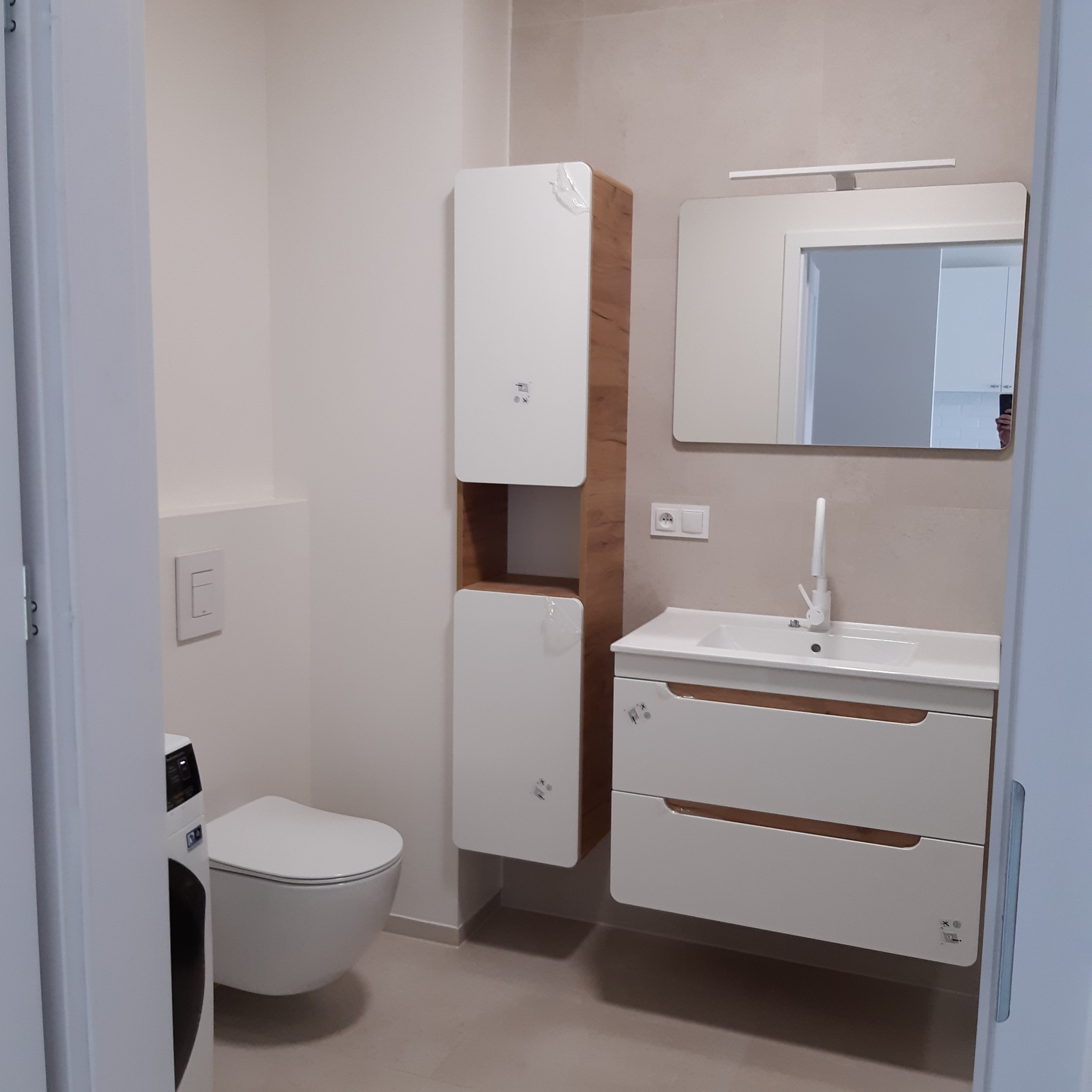 Nowoczesna łazienka z wiszącą toaletą, szafką umywalkową z szufladami i wysoką szafką, elementy zabezpieczone folią ochronną, widoczne gniazdko elektryczne i włącznik światła.