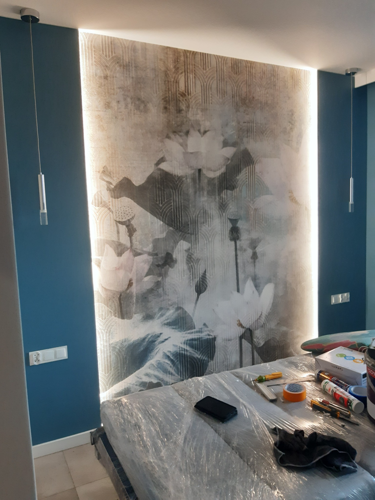 Wykończona ściana z dekoracyjną tapetą w kwiaty i geometryczne wzory, oświetlona pionowymi pasami LED po bokach, z widocznymi narzędziami i materiałami przygotowanymi do dalszych prac...