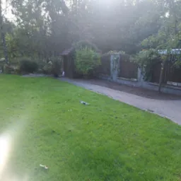 Rozległy trawnik z kostką brukową, fragmentem ogrodzenia z cegły i drewna, oraz altaną porośniętą zielenią w tle, z efektem flary słonecznej.