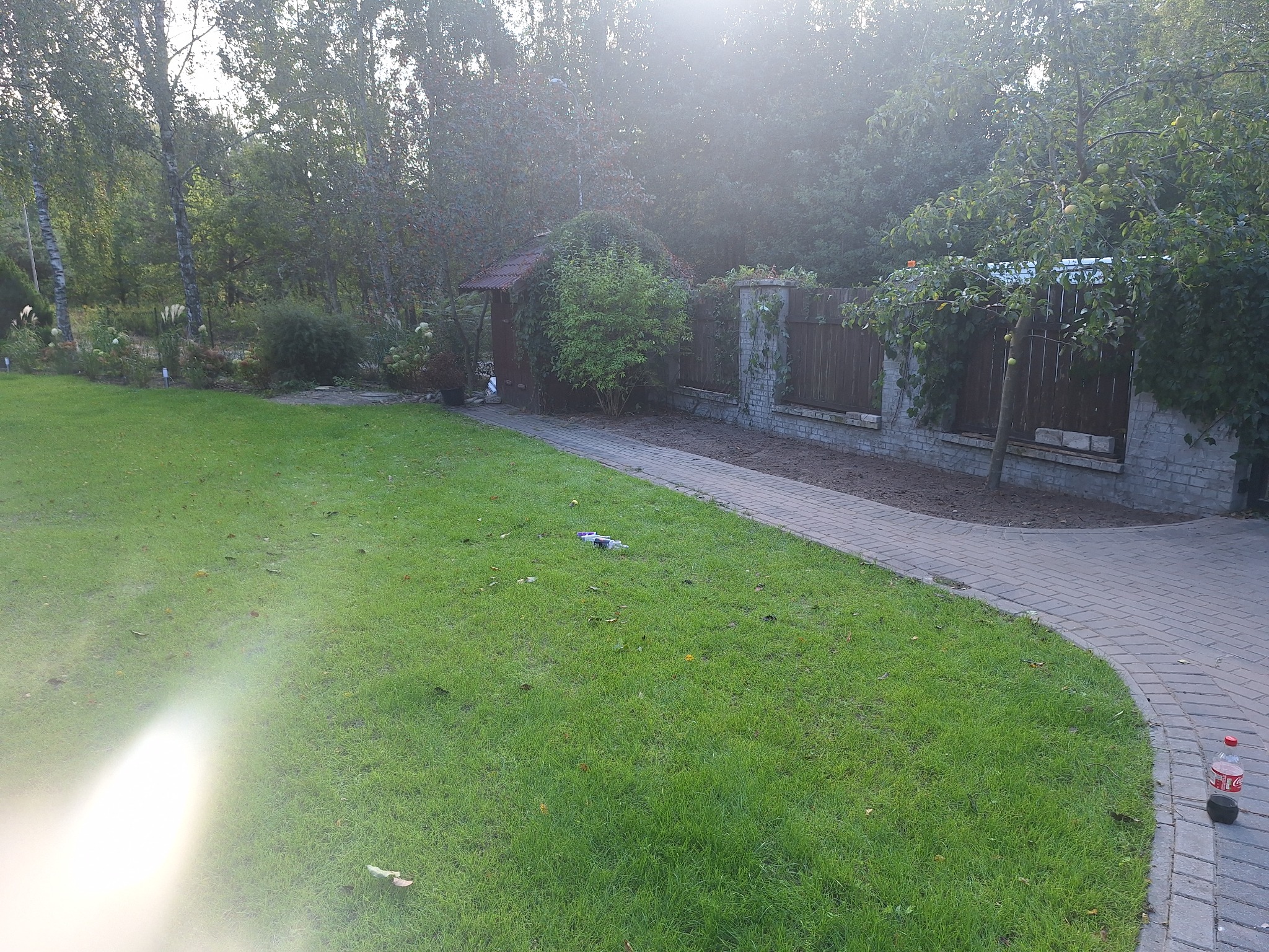 Rozległy trawnik z kostką brukową, fragmentem ogrodzenia z cegły i drewna, oraz altaną porośniętą zielenią w tle, z efektem flary słonecznej.