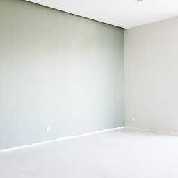 MalerGo - Pomalowane wnętrze pokoju z jedną ścianą w odcieniu pistacjowej zieleni, a pozostałe w jasnym szarym kolorze. Podłoga zabezpieczona folią malarską, widoczne gniazdka elektryczne.