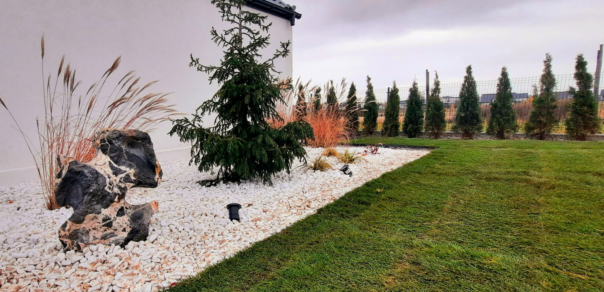 Aranżacja ogrodu z użyciem białych kamieni, ozdobnych traw i iglaków, oświetlenie punktowe, trawnik z rolki.