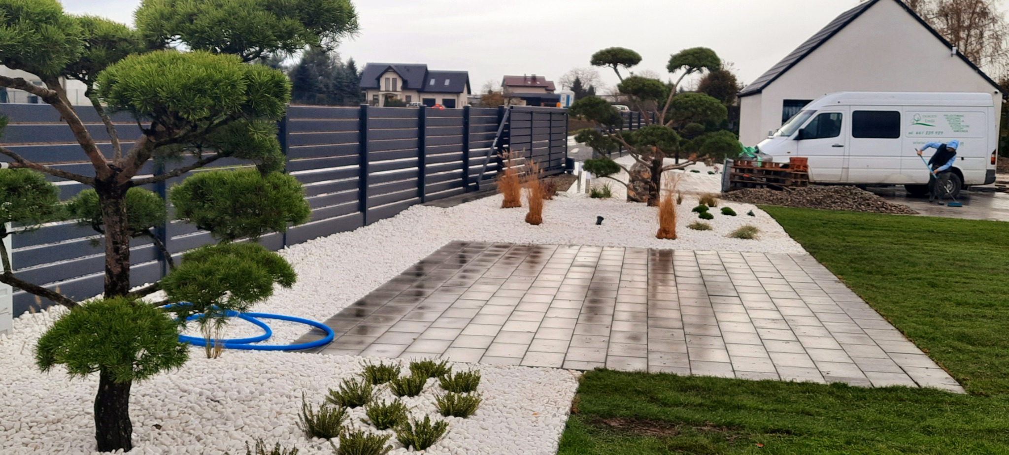 Nowoczesny ogród z wyłożonym kostką placem, ozdobnymi drzewami formowanymi, białymi kamykami i szarą palisadą w tle, z widocznym samochodem dostawczym firmy ogrodniczej.