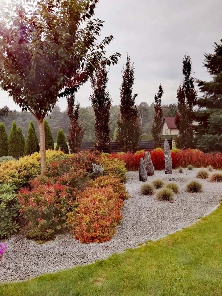 Urozmaicony ogród z drzewami, krzewami o zróżnicowanej kolorystyce i kamiennym podłożem, uzupełniony o pionowe kamienne elementy dekoracyjne.