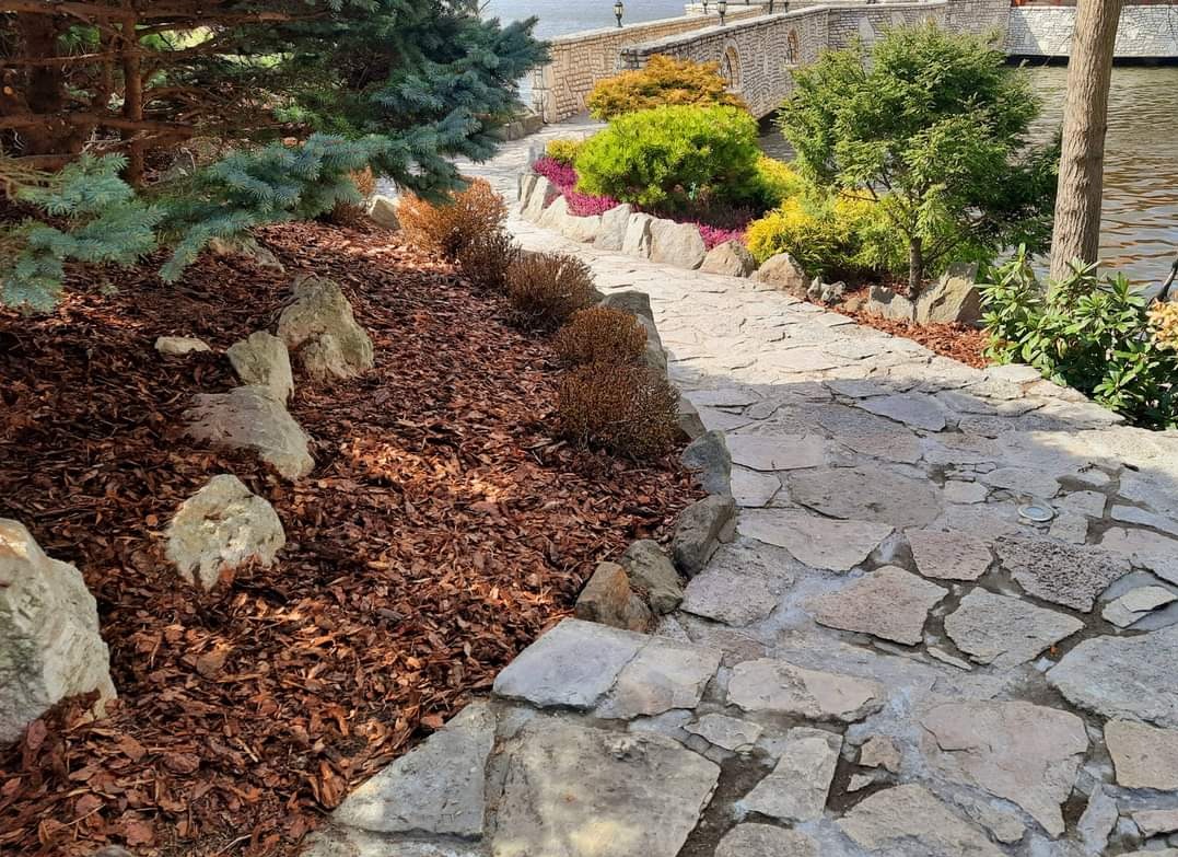 Kamienna ścieżka w ogrodzie z korą rozsypaną obok, prowadząca w stronę mostu nad wodą, otoczona różnorodną roślinnością.