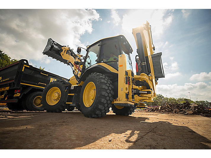 Żółta koparko-ładowarka Caterpillar z podniesioną łyżką i ramieniem kopiącym, stojąca obok czarnej wywrotki na placu budowy, widok z dołu, silne słońce.