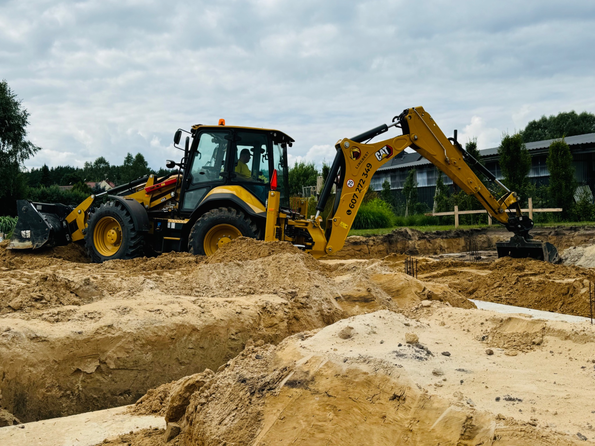 Żółta koparko-ładowarka CAT w trakcie wykopu pod fundamenty na piaszczystym terenie, widoczne zbrojenia i operator w kabinie.