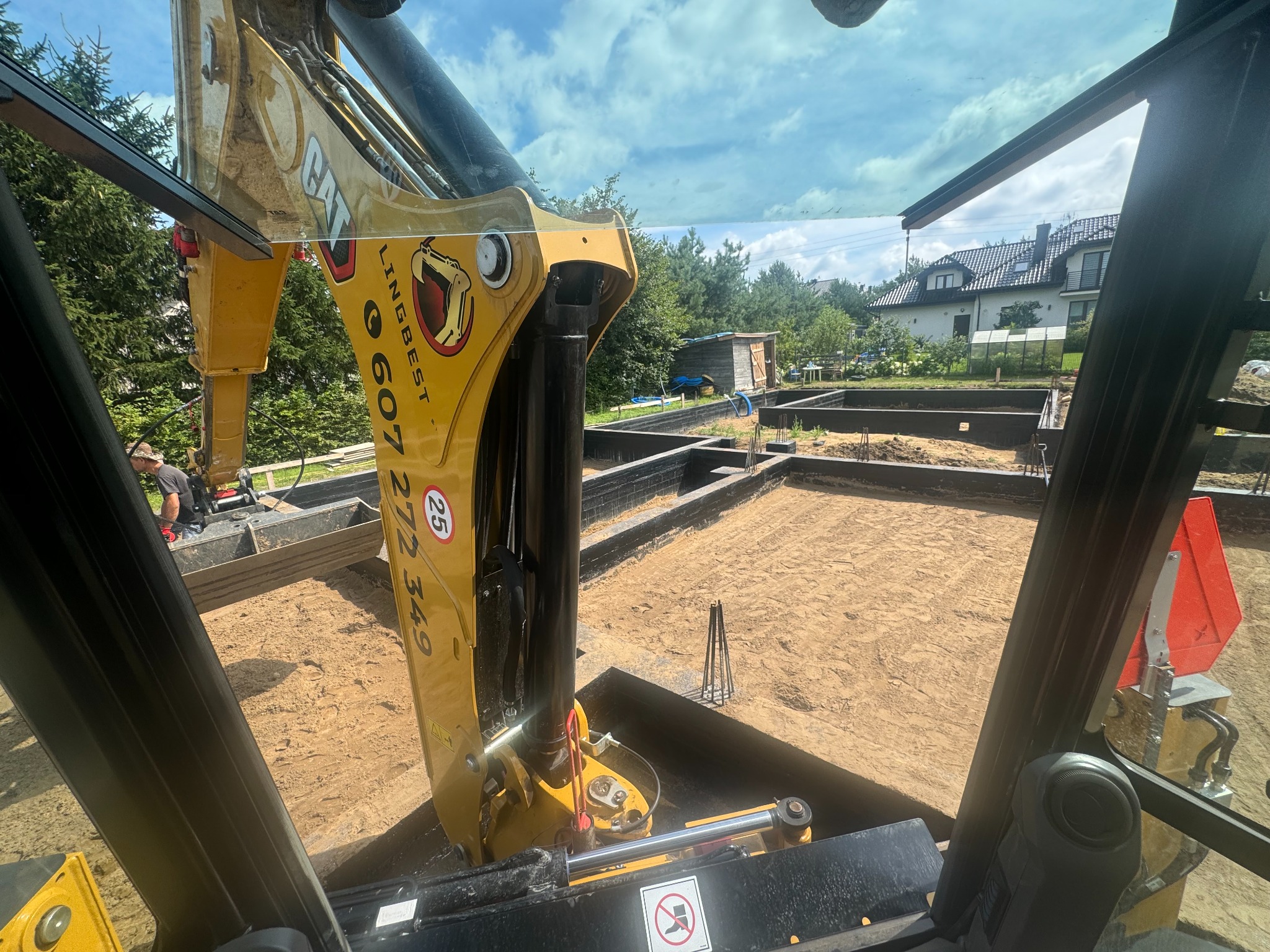 Widok z kabiny koparki CAT na wykop pod fundamenty domu jednorodzinnego, widoczne zbrojenia i przygotowany teren, w tle dom i zieleń.