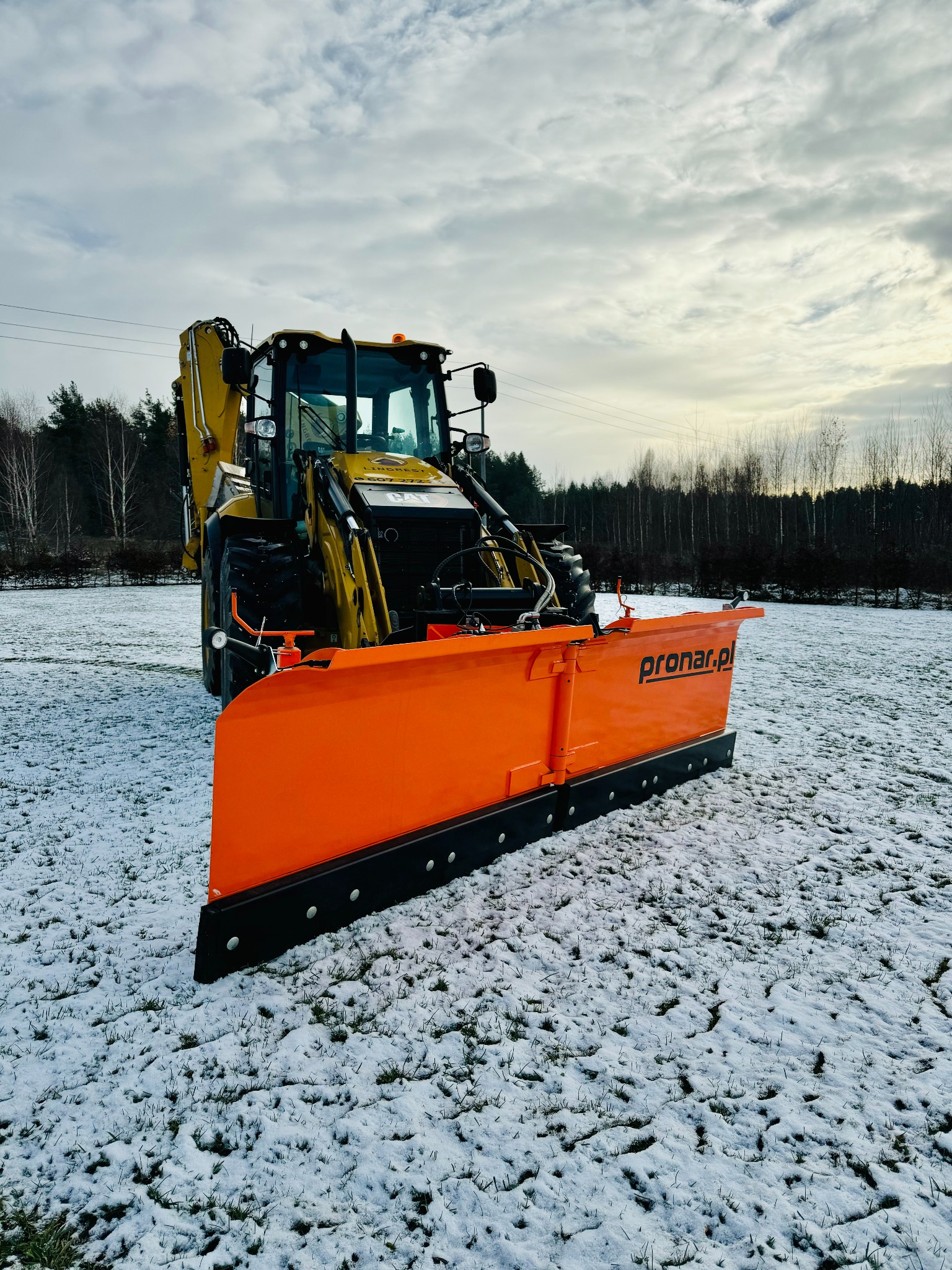 Żółta ładowarka teleskopowa z pomarańczowym pługiem śnieżnym Pronar na zaśnieżonym polu, widok z przodu, pochmurne niebo.