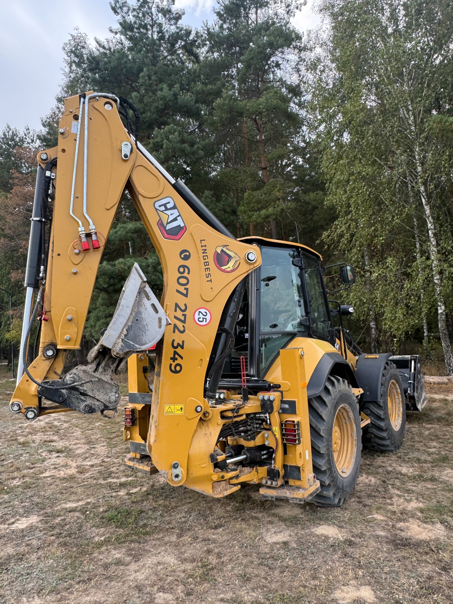 Żółta koparko-ładowarka CAT zaparkowana na piaszczystym terenie z lasem w tle, widoczne ślady błota na łyżce i oponach, numer telefonu na ramieniu koparki.