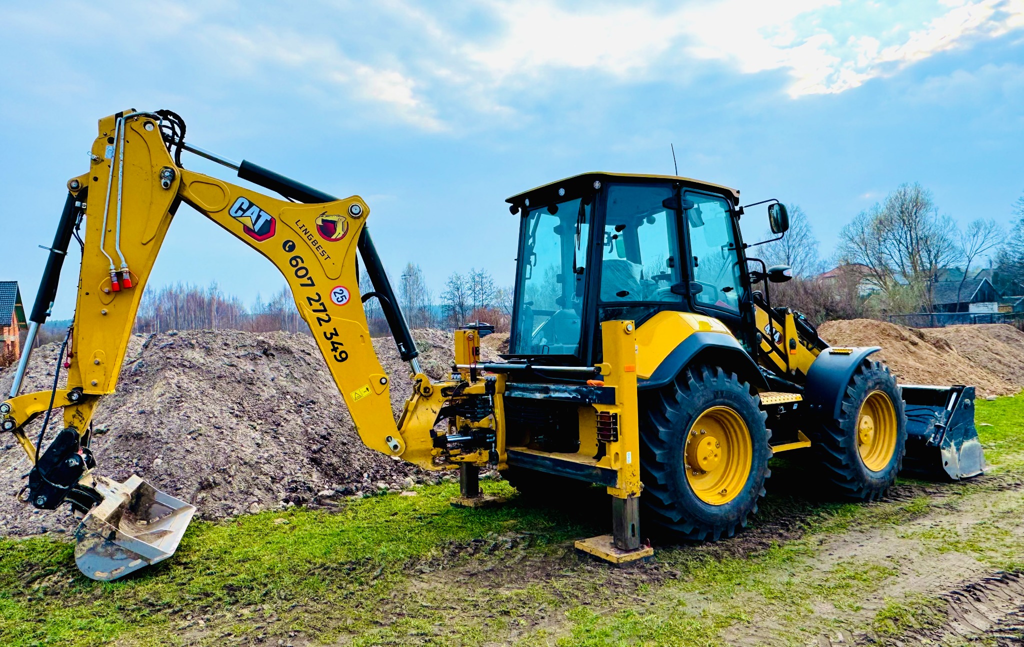 Żółta koparko-ładowarka CAT z wysuniętymi podporami stabilizującymi, na trawiastym terenie z pryzmą ziemi w tle, Białystok.