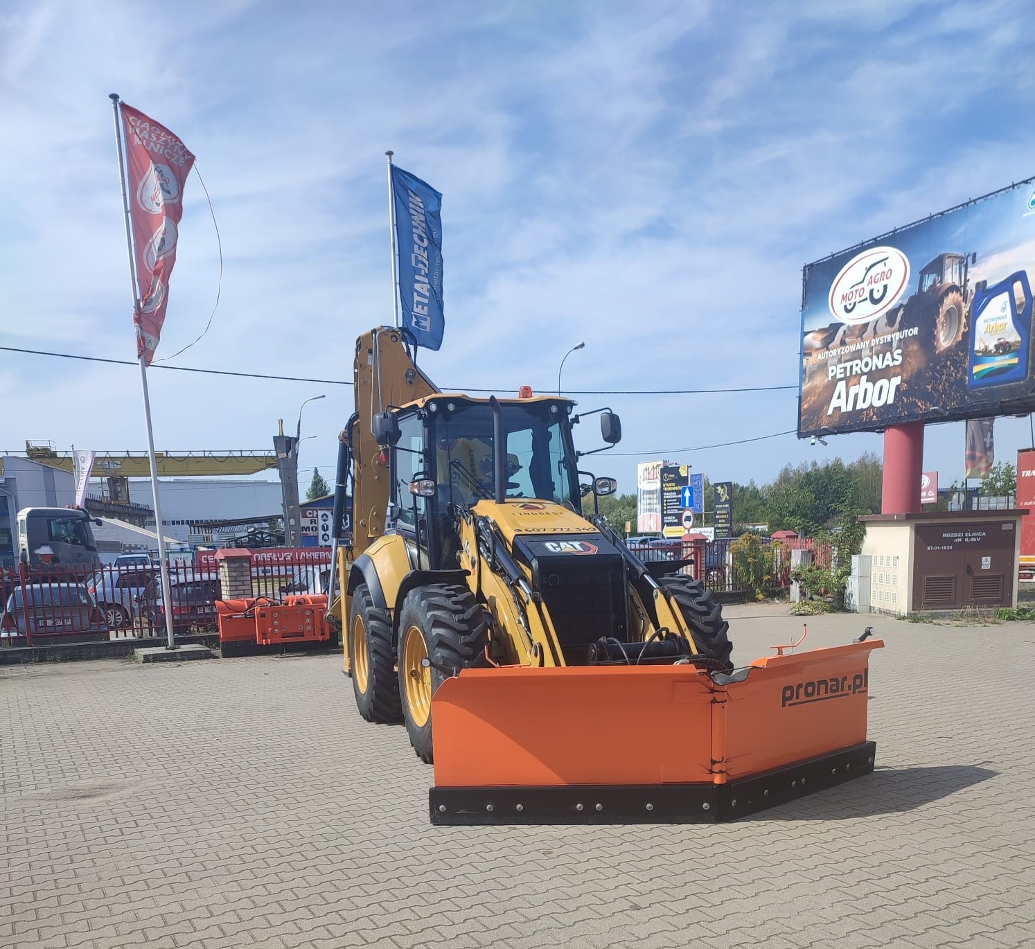 Żółta koparko-ładowarka CAT z pomarańczowym pługiem Pronar na kostce brukowej, na tle billboard z reklamą olejów silnikowych i flagi.