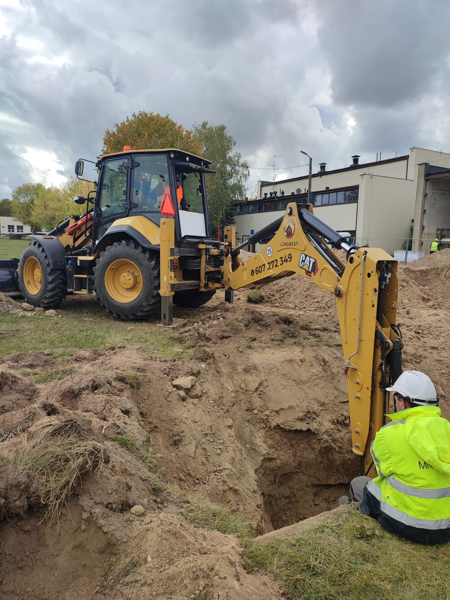 Żółta koparko-ładowarka CAT przy wykopie, obok pracownik w kasku i kamizelce. Widoczne budynki w tle i pochmurne niebo. Prace ziemne w toku.