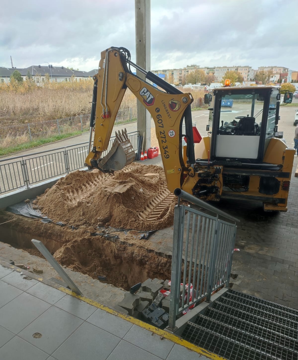 Żółta koparko-ładowarka CAT przy wykopie w płytkach chodnikowych, z hałdą piasku i widocznymi śladami opon. W tle budynki mieszkalne i pochmurne niebo.