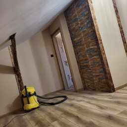 Agnieszka bortnowska - Poddasze po remoncie z drewnianymi belkami, ścianą z kamienia dekoracyjnego i odkurzaczem na podłodze z paneli.