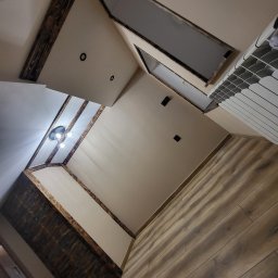 Agnieszka bortnowska - Wykończone wnętrze poddasza z drewnianymi belkami stropowymi, widoczne oświetlenie, grzejnik i drewniana podłoga.