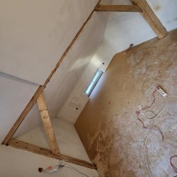 Agnieszka bortnowska - Poddasze w trakcie prac w Siemiatyczach: widoczne drewniane elementy konstrukcyjne, ściany po nałożeniu gładzi, okno, oraz rozciągnięte kable elektryczne i przedłużacz na podłodze.