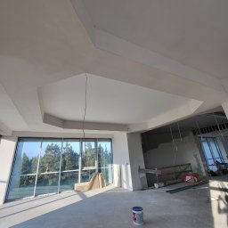 Agnieszka bortnowska - Wnętrze w trakcie prac wykończeniowych: sufit z elementami dekoracyjnymi, duże okno z widokiem na drzewa, puszka farby na podłodze, widoczne elementy konstrukcyjne i instalacje.