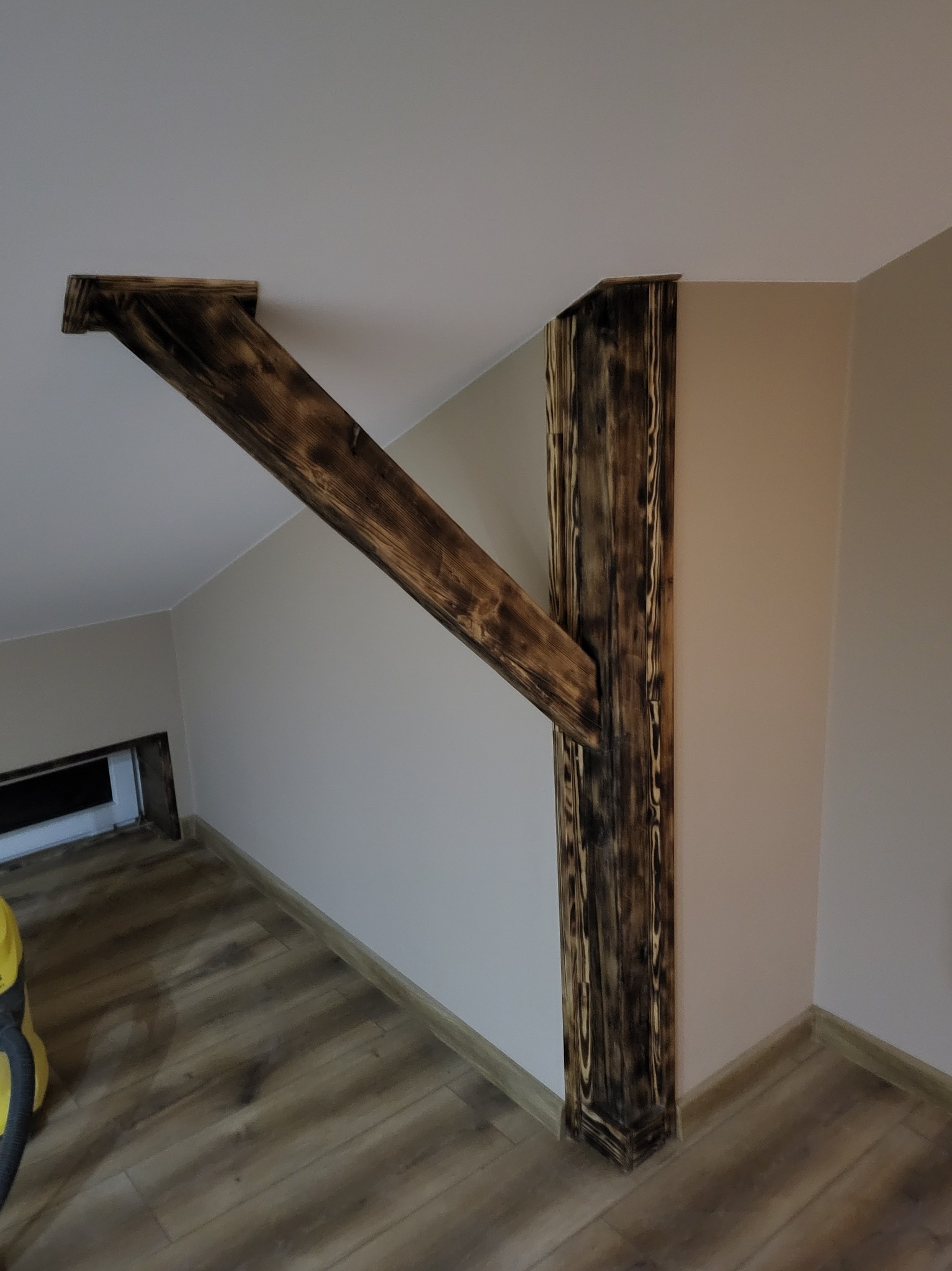 Drewniana konstrukcja nośna z belek o wypalanej strukturze, wzmacniająca poddasze, widoczna na tle ściany pomalowanej na dwa odcienie beżu.