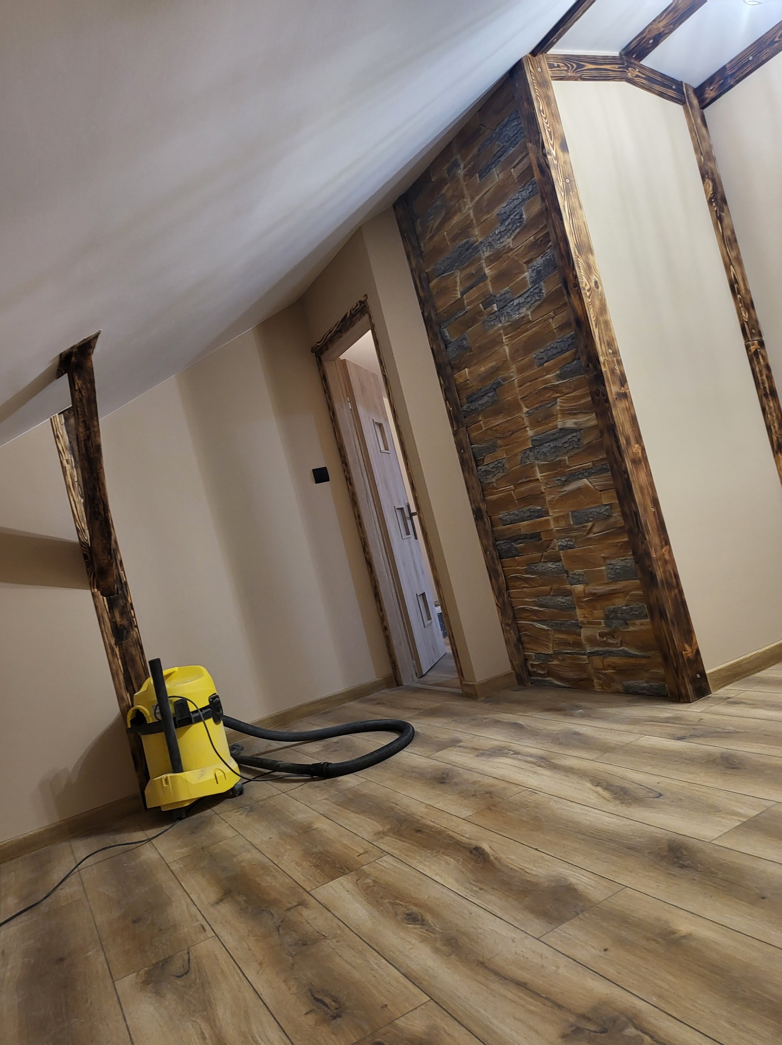 Poddasze po remoncie z drewnianymi belkami, ścianą z kamienia dekoracyjnego i odkurzaczem na podłodze z paneli.