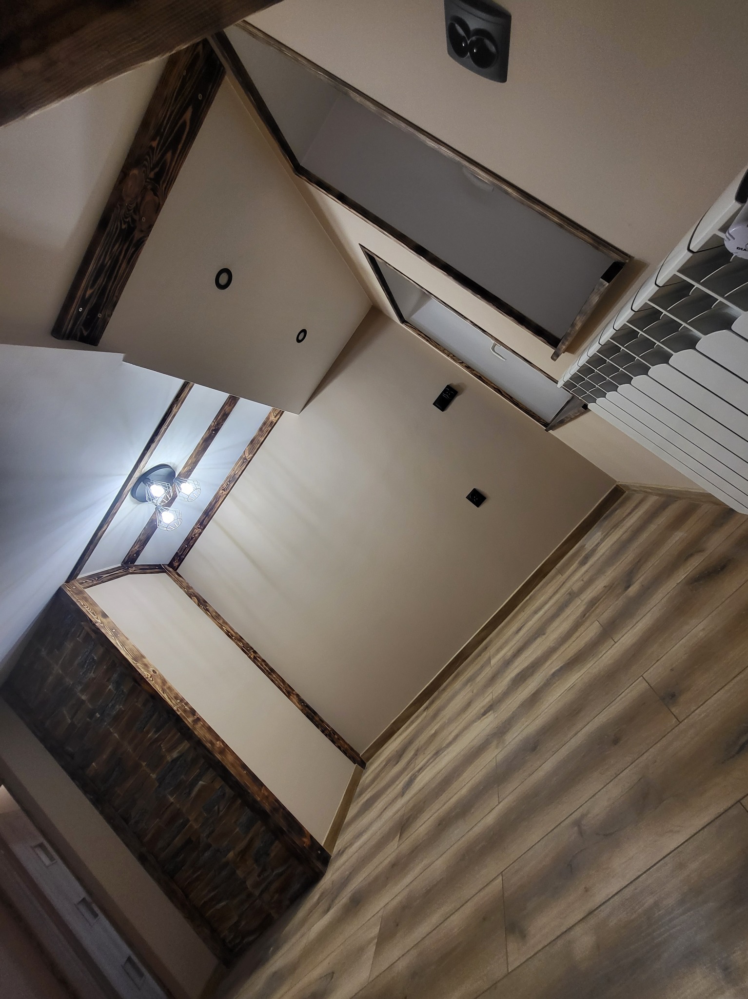 Wykończone wnętrze poddasza z drewnianymi belkami stropowymi, widoczne oświetlenie, grzejnik i drewniana podłoga.