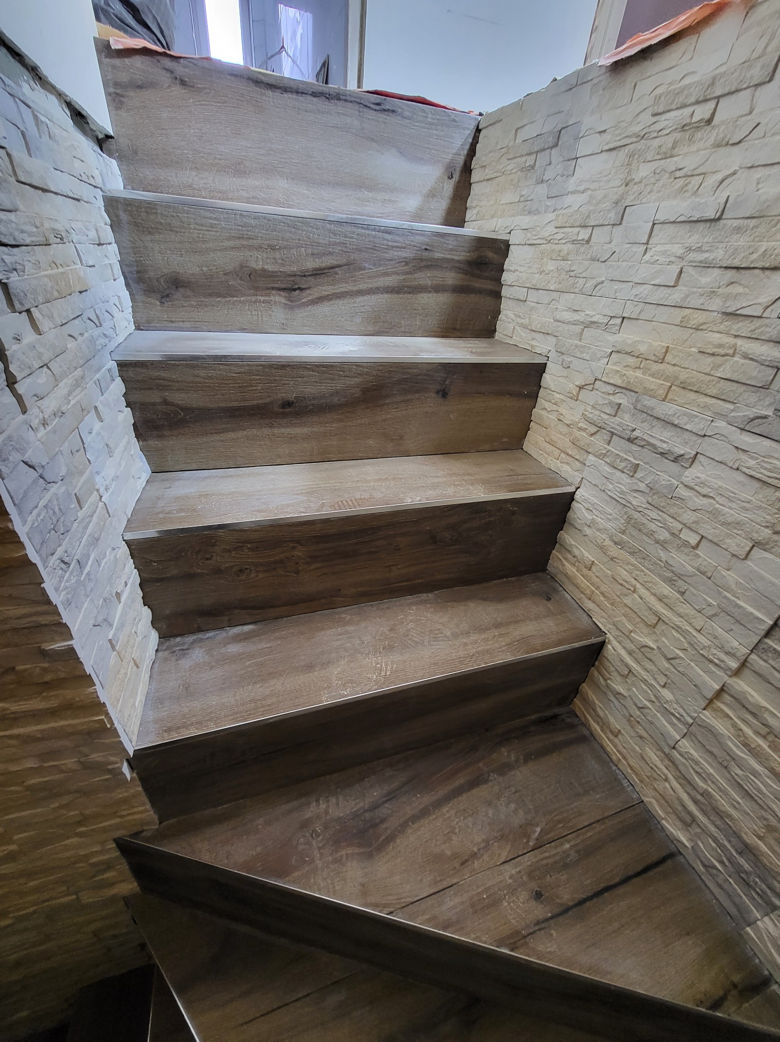 Schody wykończone panelami drewnopodobnymi, ściana obłożona dekoracyjnym kamieniem w odcieniach beżu, widok z dołu.