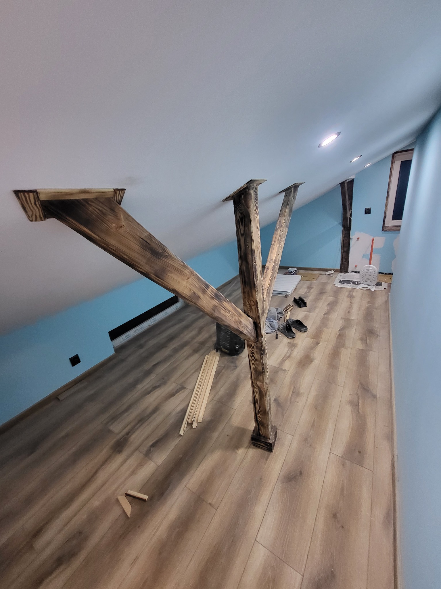 Drewniane belki konstrukcyjne w skośnym pokoju na poddaszu, jasne panele podłogowe, błękitne ściany, widoczne prace wykończeniowe.