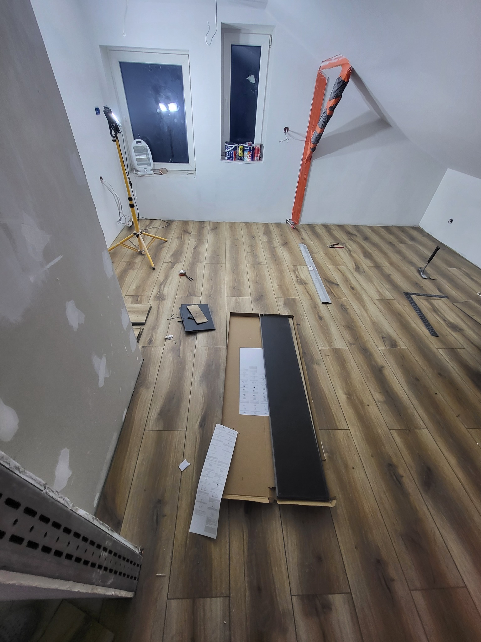 Poddasze w trakcie układania paneli podłogowych, widoczne narzędzia, lampa budowlana i częściowo wykończona ściana z widocznymi miejscami po szpachlowaniu.