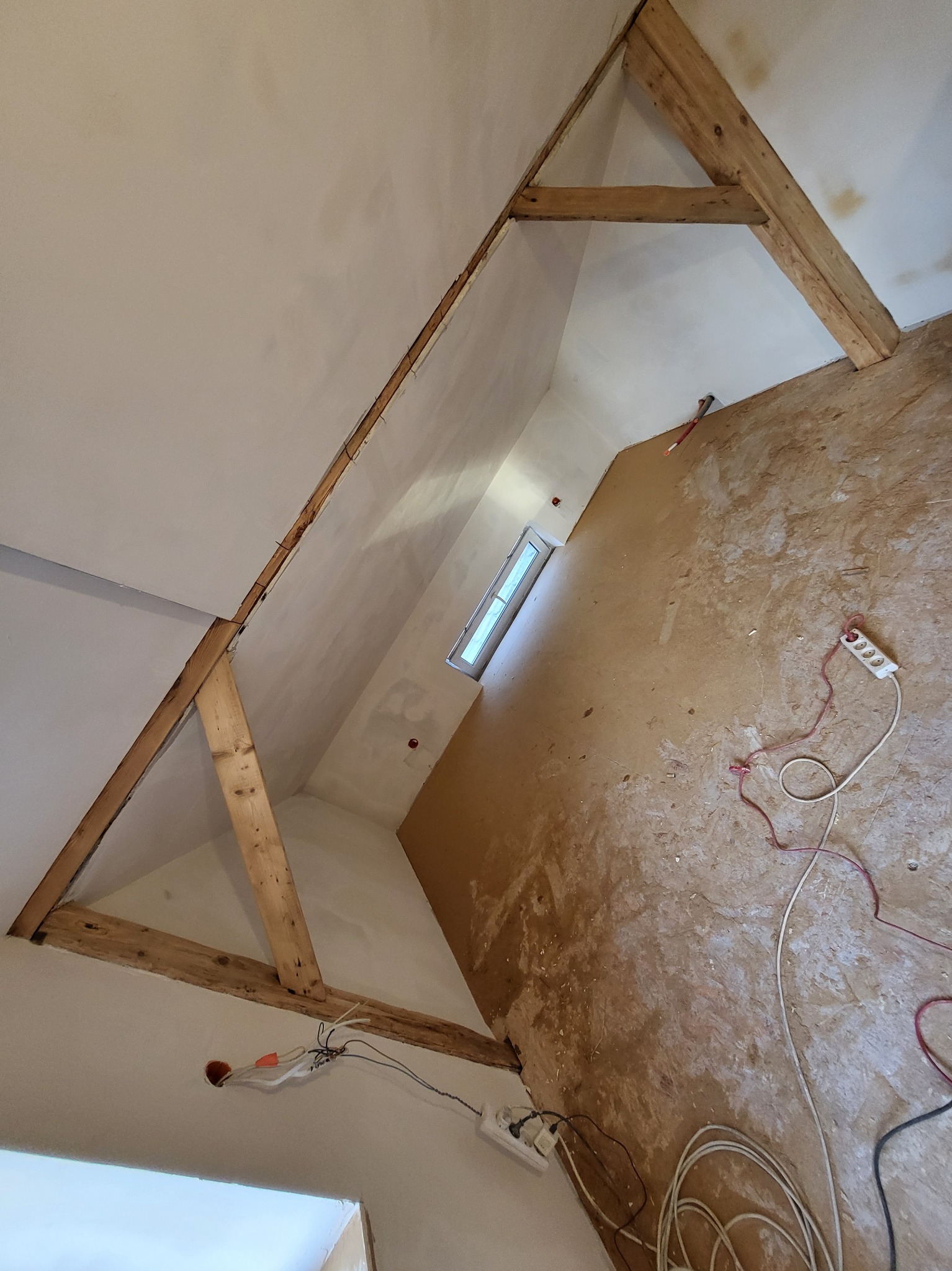 Poddasze w trakcie prac w Siemiatyczach: widoczne drewniane elementy konstrukcyjne, ściany po nałożeniu gładzi, okno, oraz rozciągnięte kable elektryczne i przedłużacz na podłodze.