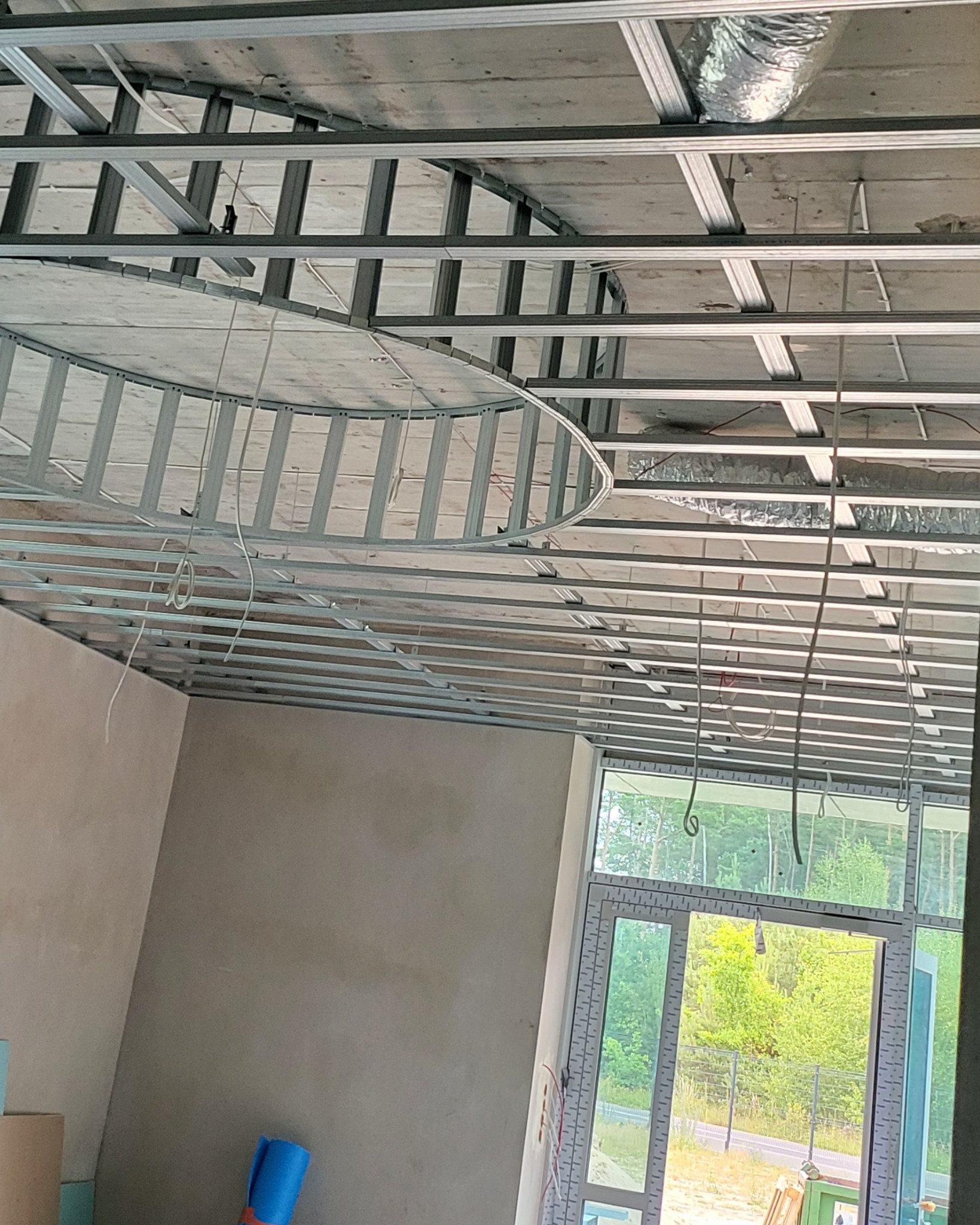 Metalowa konstrukcja sufitu podwieszanego z elementem owalnym, widoczne przewody i surowa ściana przed wykończeniem.