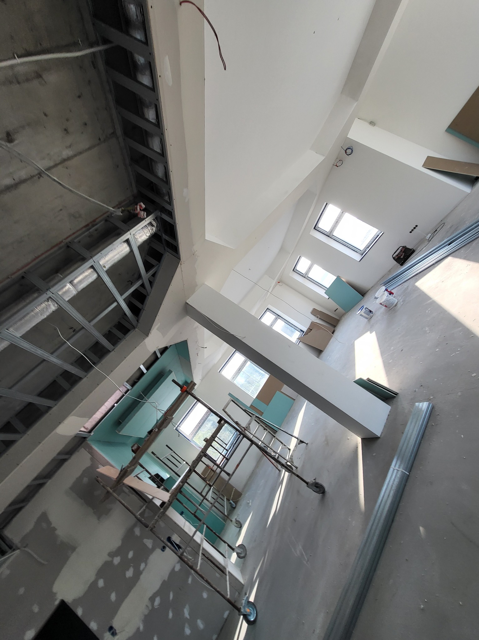 Wnętrze w trakcie prac wykończeniowych, widoczne metalowe profile sufitowe, rusztowanie, płyty kartonowo-gipsowe oraz częściowo wygładzone ściany. W tle okna dachowe wpuszczające dużo światła.