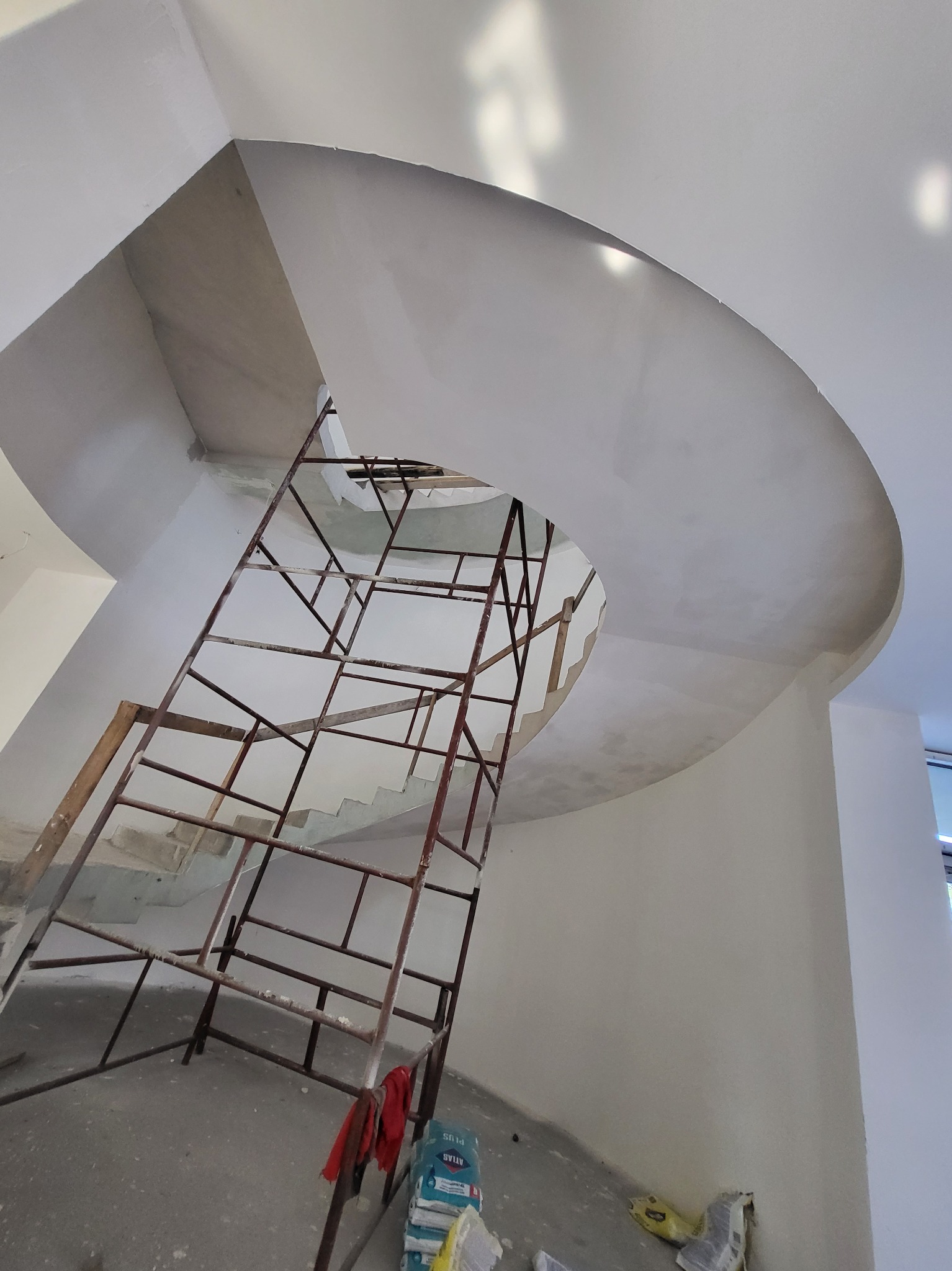 Klatka schodowa w trakcie wykańczania, widoczne rusztowanie, surowe ściany i strop z zaokrąglonymi krawędziami, worki z materiałami budowlanymi na podłodze.