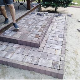 złota rączka - Ułożona kostka brukowa tworząca schody wejściowe na taras, widoczne fragmenty osoby w szortach i tenisówkach, piasek wokół nowej konstrukcji.