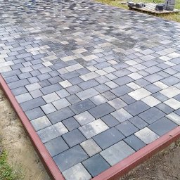 złota rączka - Ułożona kostka brukowa w odcieniach szarości i grafitu, obramowana czerwonobrązową metalową listwą. Częściowo mokra powierzchnia odbija światło, w tle paleta z elementami kostki.