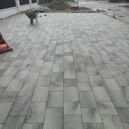 złota rączka - Ułożona kostka brukowa w odcieniach szarości na podjeździe, widoczny wózek budowlany, ubijarka, oraz przygotowany stos materiałów, w tle ulica i zabudowa jednorodzinna.